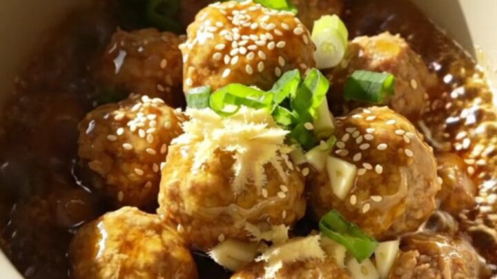A close-up of golden-amber glazed meatballs in a ceramic slow cooker, garnished with green scallions.