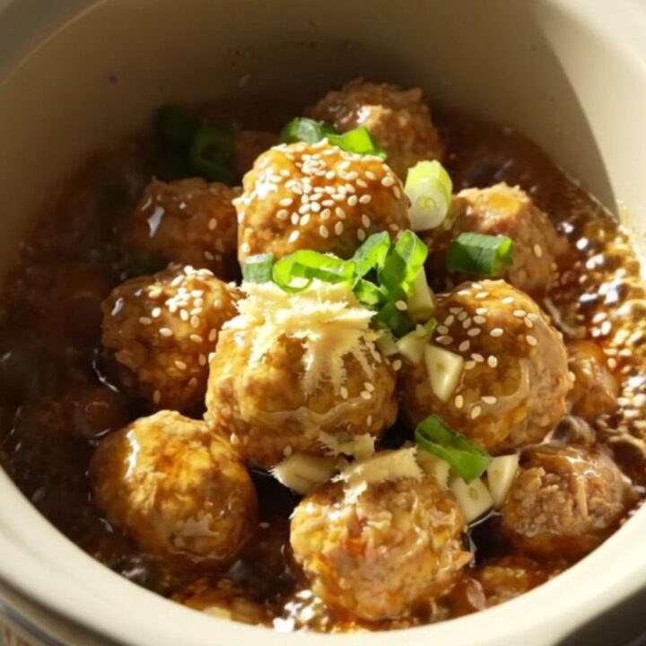 A close-up of golden-amber glazed meatballs in a ceramic slow cooker, garnished with green scallions.