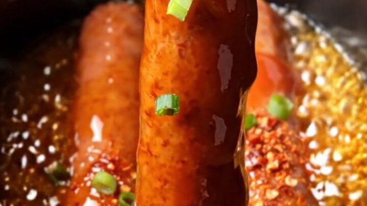 A close-up of glistening, deep-red glazed Lil' Smokies in a black ceramic slow cooker, garnished with green scallions.