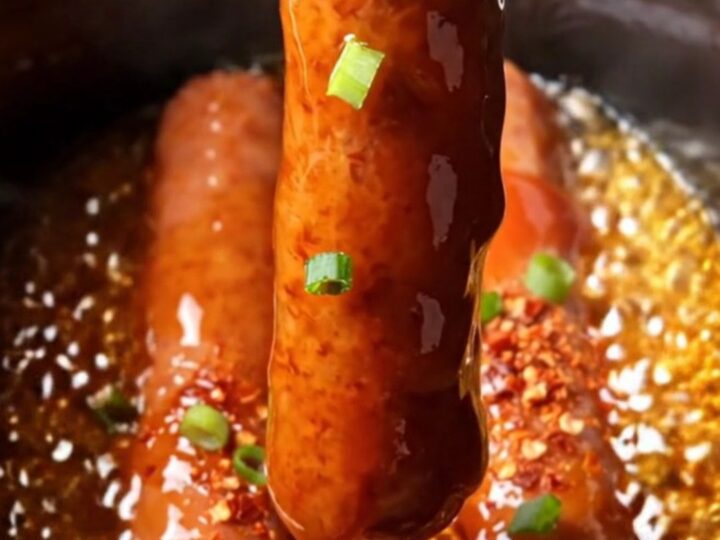A close-up of glistening, deep-red glazed Lil' Smokies in a black ceramic slow cooker, garnished with green scallions.