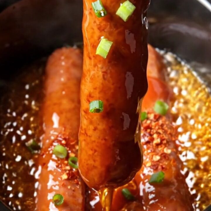 A close-up of glistening, deep-red glazed Lil' Smokies in a black ceramic slow cooker, garnished with green scallions.
