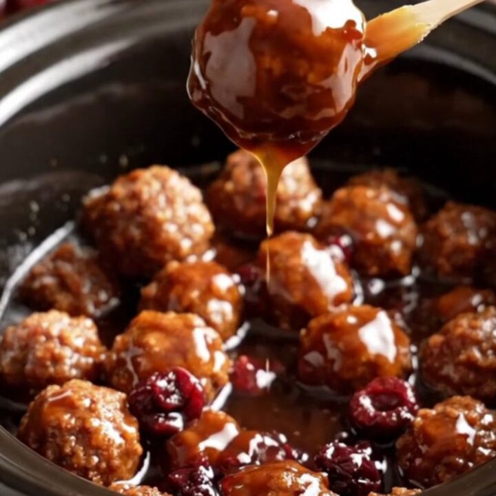 A close-up of glossy, dark mahogany-glazed meatballs in a cherry-red ceramic slow cooker.