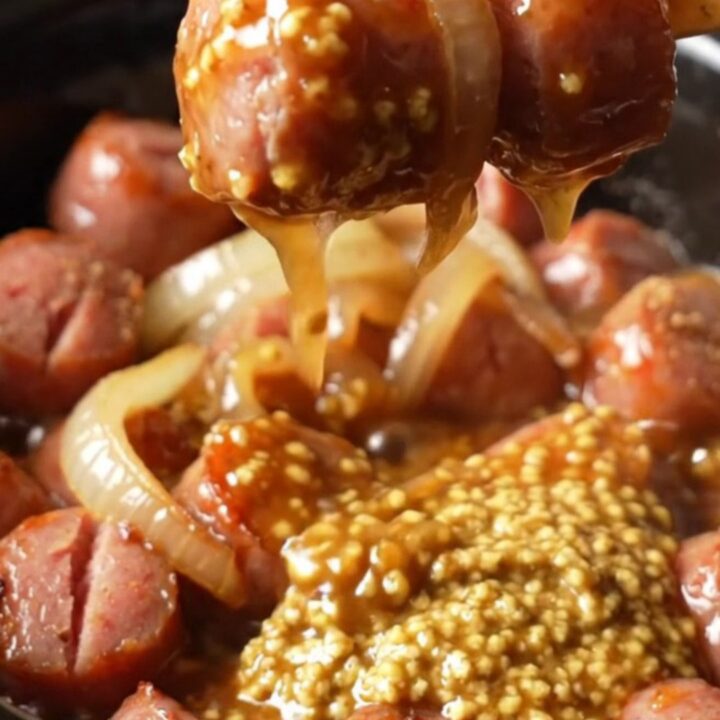 A close-up shot of bratwurst coins in a rich, glossy mustard glaze inside a rustic stoneware slow cooker.