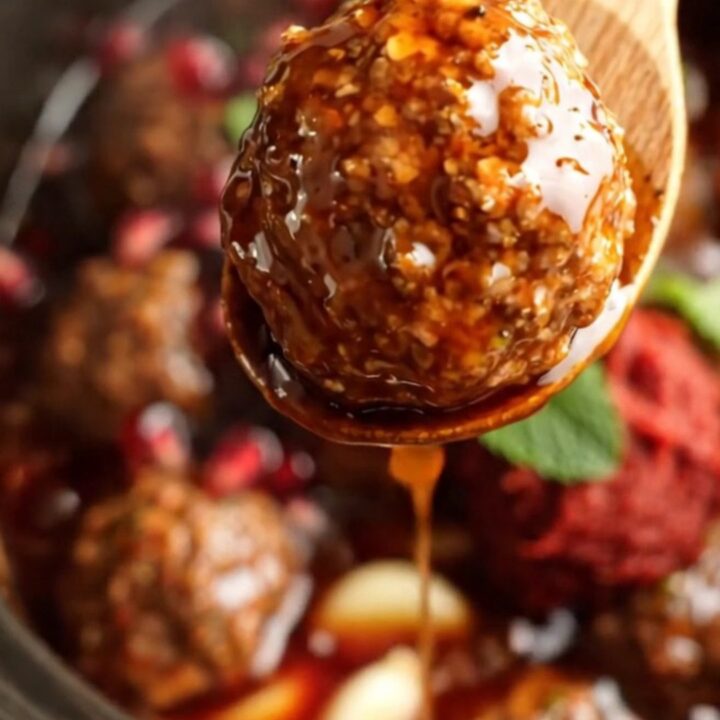 A close-up of pomegranate and harissa glazed lamb meatballs in a terracotta crockpot, garnished with fresh mint and arils.
