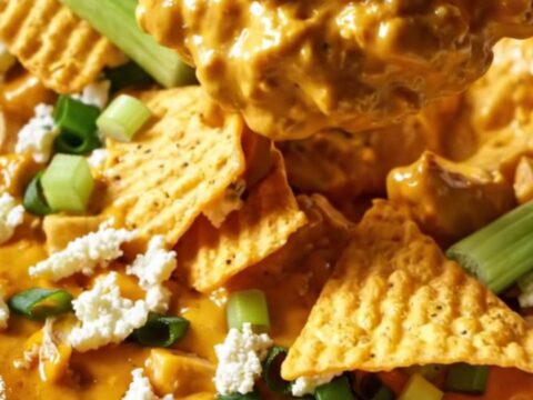 A close-up of a ridge-cut tortilla chip being dipped into a creamy, bubbling buffalo chicken dip in a white ceramic crockpot.