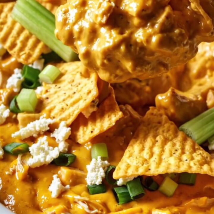 A close-up of a ridge-cut tortilla chip being dipped into a creamy, bubbling buffalo chicken dip in a white ceramic crockpot.