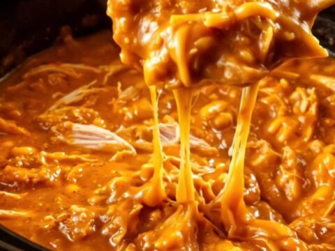 A close-up of a pita chip being dipped into a creamy, bubbling honey sriracha chicken dip in a black crockpot.