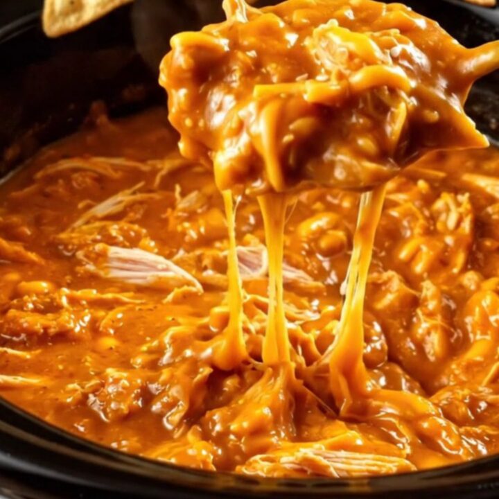 A close-up of a pita chip being dipped into a creamy, bubbling honey sriracha chicken dip in a black crockpot.