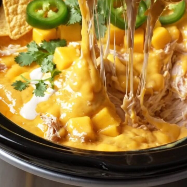 A close-up view of a lime tortilla chip plunging into a creamy, orange-gold Crockpot chicken dip with visible mango chunks.