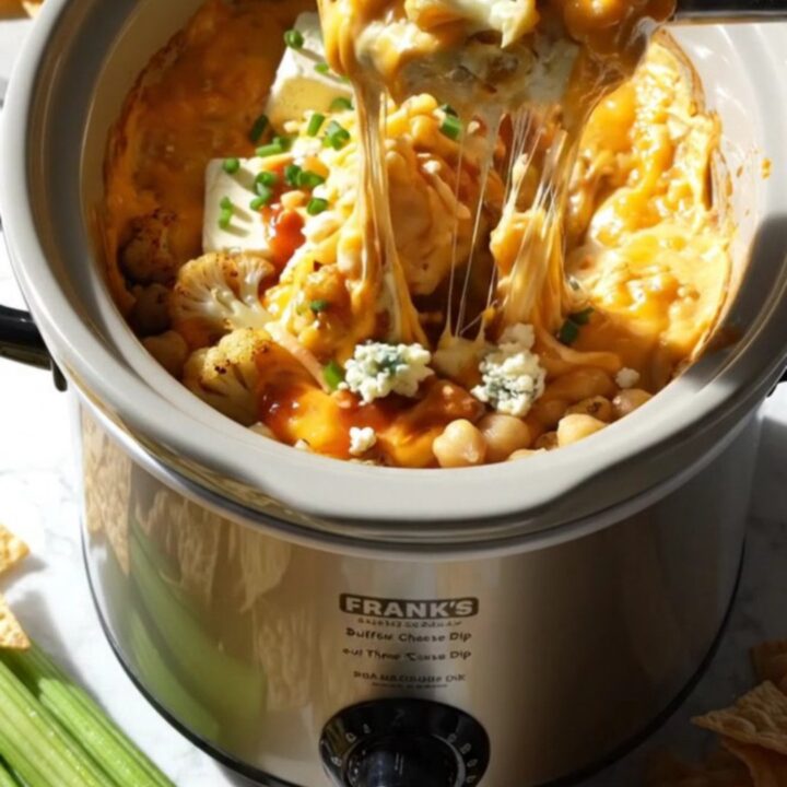 A close-up shot of a pita chip being dipped into a bubbling, cheesy buffalo cauliflower dip in a slow cooker.