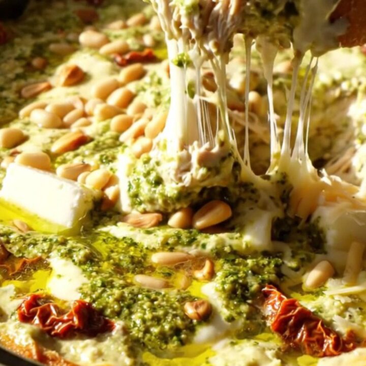 A close-up of a rustic ceramic slow cooker filled with creamy pesto chicken dip, topped with pine nuts and fresh herbs.