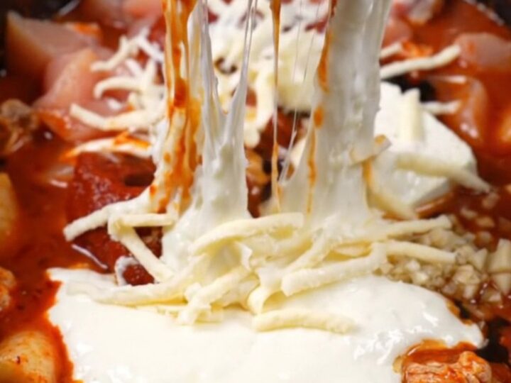 A close-up of a spicy Korean chicken dip in a black ceramic pot with a massive mozzarella cheese pull.