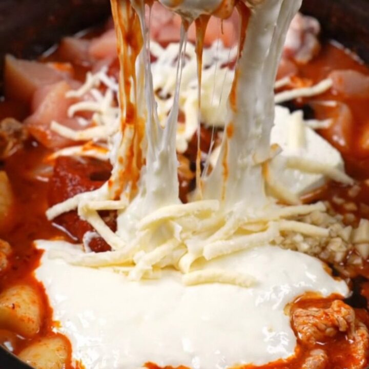 A close-up of a spicy Korean chicken dip in a black ceramic pot with a massive mozzarella cheese pull.
