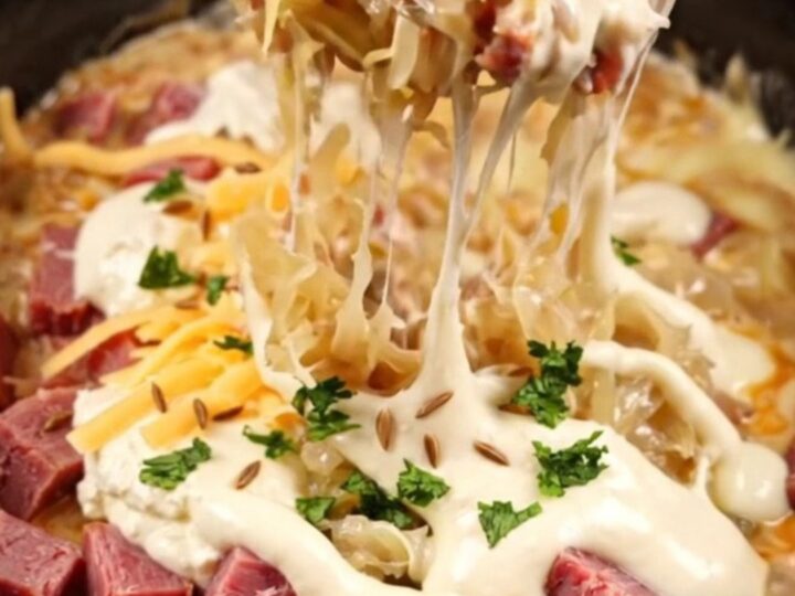 A close-up of a bubbling, creamy Reuben dip in a dark stoneware crockpot with a piece of toasted marbled rye bread.