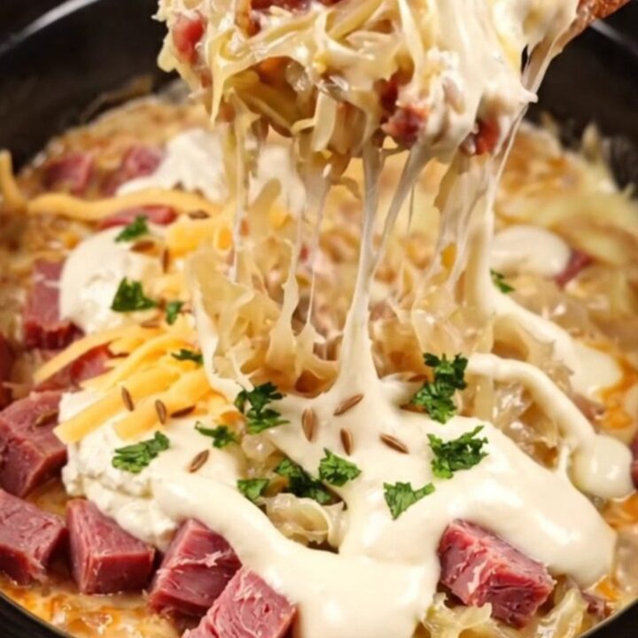 A close-up of a bubbling, creamy Reuben dip in a dark stoneware crockpot with a piece of toasted marbled rye bread.