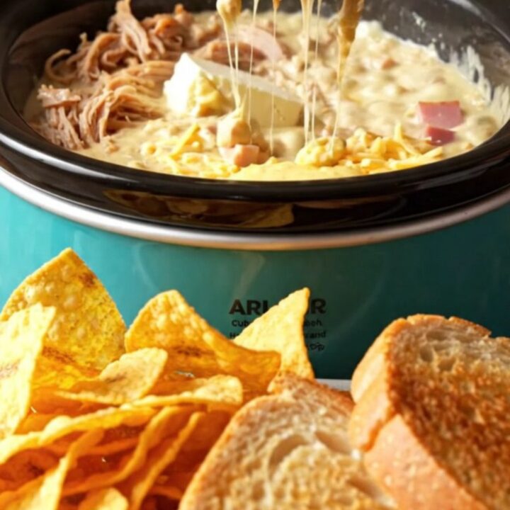 A close-up of a warm, creamy Cuban sandwich dip in a turquoise ceramic crockpot, scooped with a crispy plantain chip.