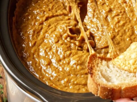 A close-up shot of a toasted baguette dipped into a bubbling, golden-brown French onion and Gruyere dip.