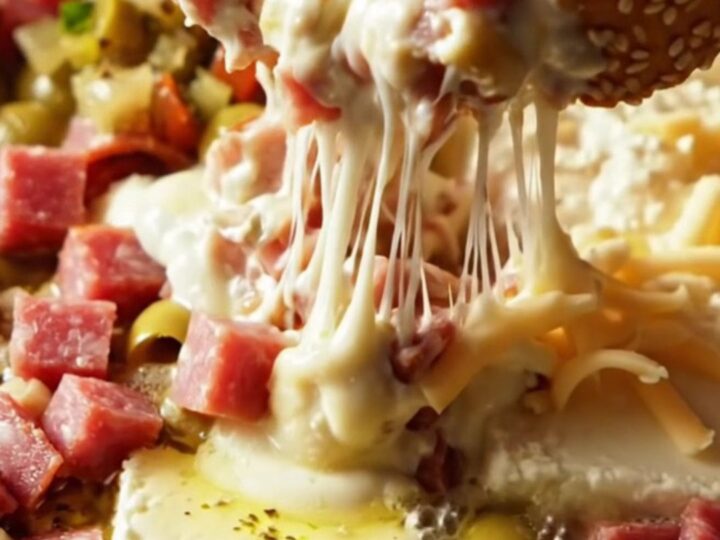 A close-up of a warm, bubbling Muffuletta dip in a white ceramic slow cooker with a long, gooey provolone cheese pull.