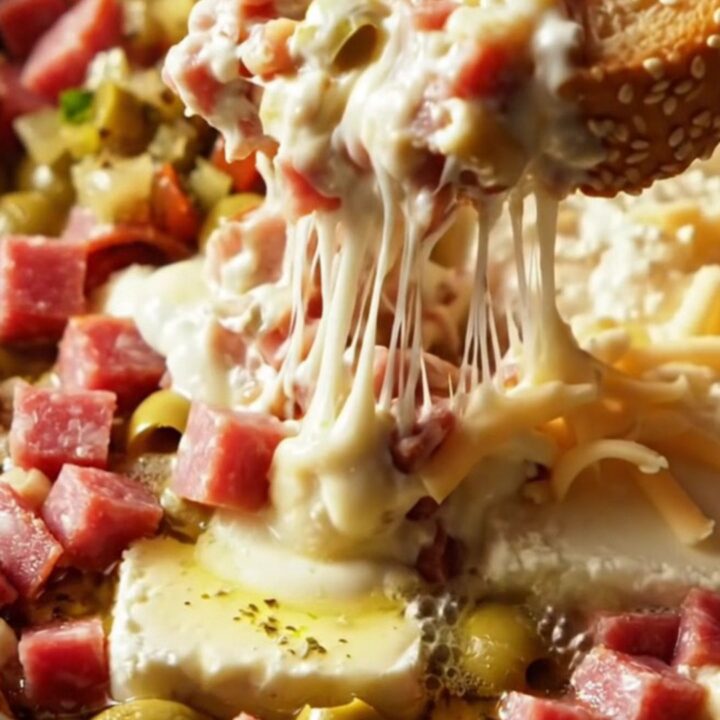 A close-up of a warm, bubbling Muffuletta dip in a white ceramic slow cooker with a long, gooey provolone cheese pull.