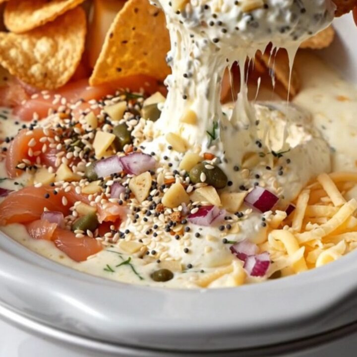 A close-up shot of a bagel chip being dipped into a creamy, bubbling smoked salmon dip inside a ceramic slow cooker.