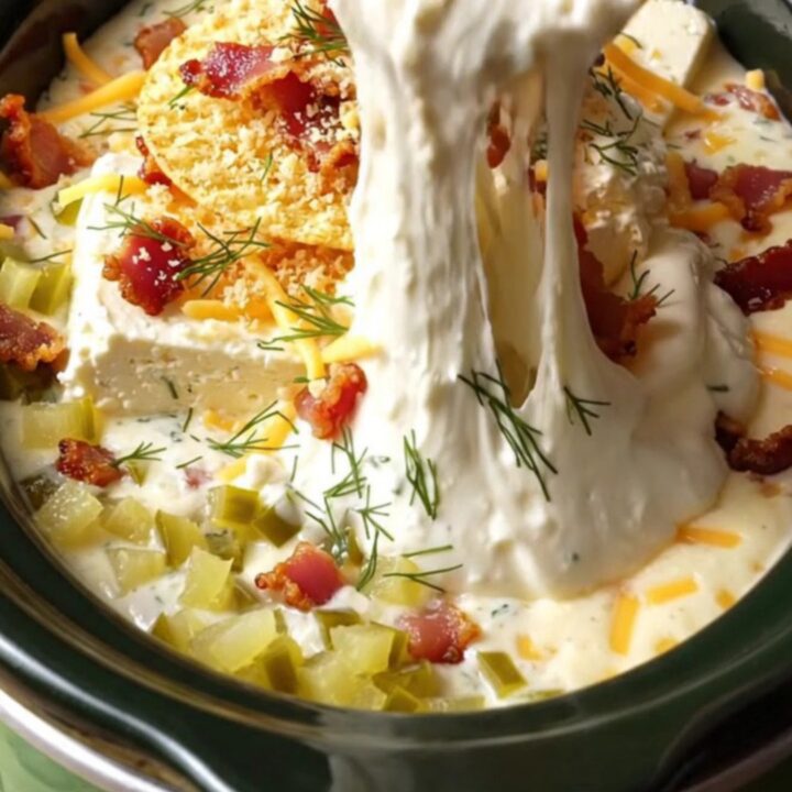 A close-up of a potato chip being dipped into a creamy, bubbling slow-cooker dip topped with bacon and panko.