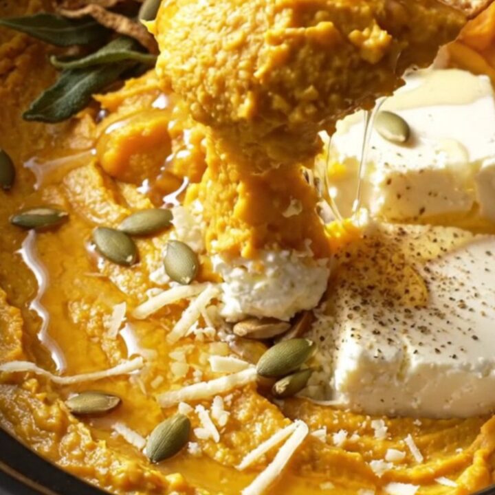 A close-up of a creamy butternut squash and goat cheese dip in a ceramic crockpot with a pita chip dipping in.