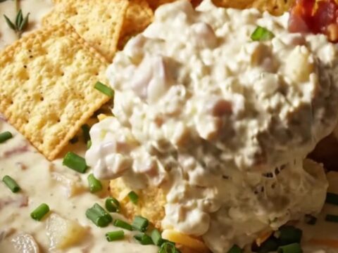 A creamy clam chowder dip in a stoneware crockpot topped with oyster crackers and chives.