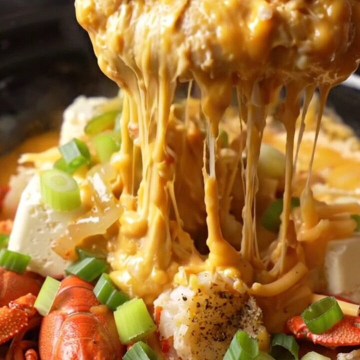 A close-up of a deep red ceramic slow cooker filled with creamy, golden-orange crawfish étouffée queso dip.