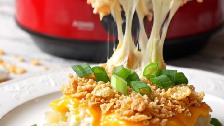 A close-up of a silver spoon scooping creamy, cheesy funeral potatoes from a red ceramic slow cooker.