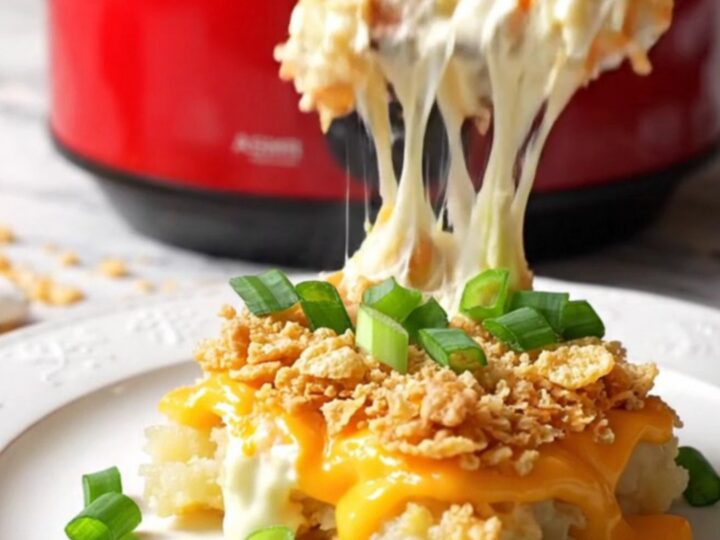 A close-up of a silver spoon scooping creamy, cheesy funeral potatoes from a red ceramic slow cooker.