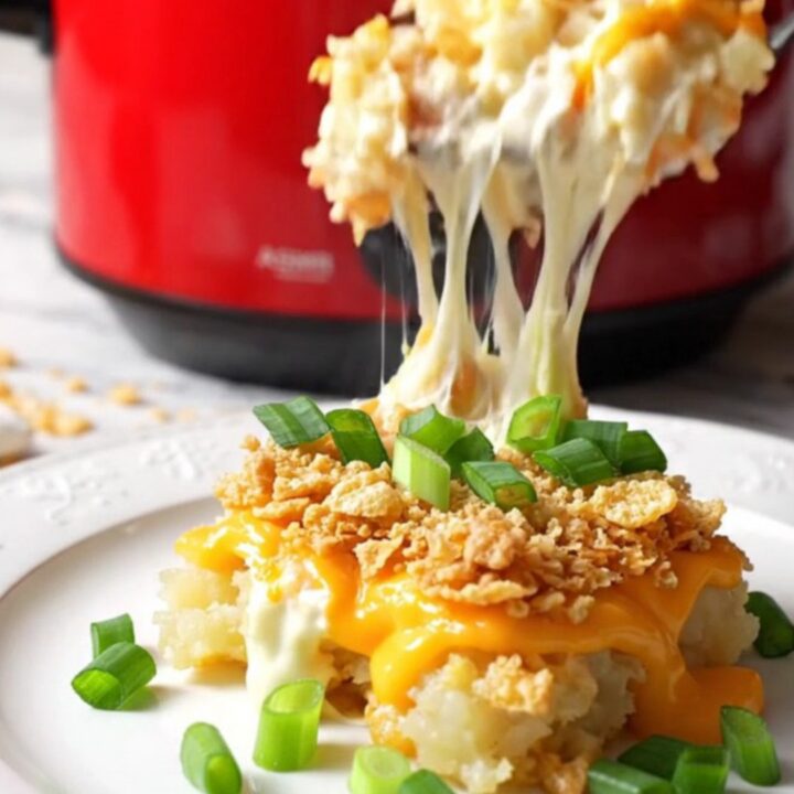 A close-up of a silver spoon scooping creamy, cheesy funeral potatoes from a red ceramic slow cooker.
