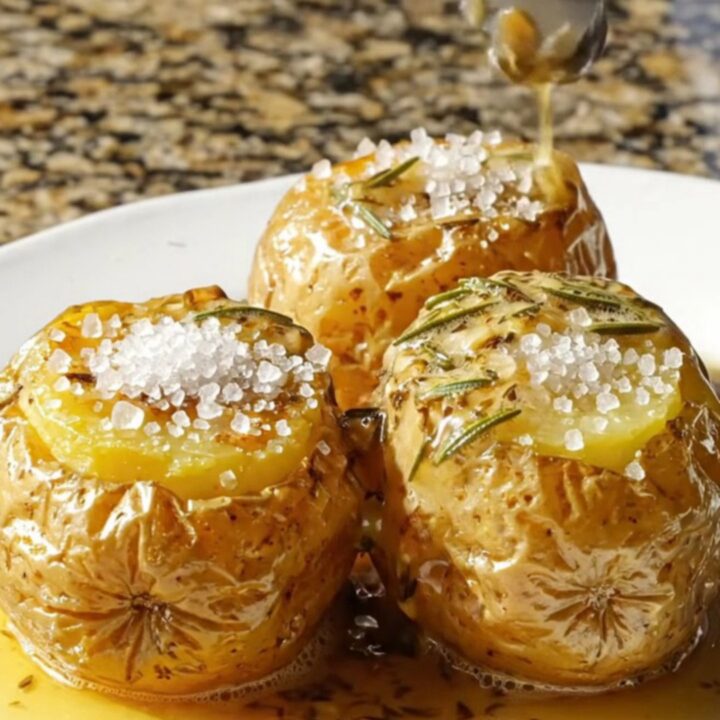 A close-up view of three perfectly cooked fondant potatoes glistening with a rich, amber herb butter glaze on a platter.