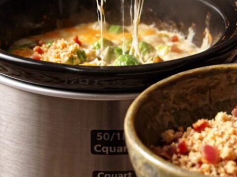 A close-up of a ceramic slow cooker filled with creamy Brussels sprouts au gratin, topped with crispy bacon and breadcrumbs.
