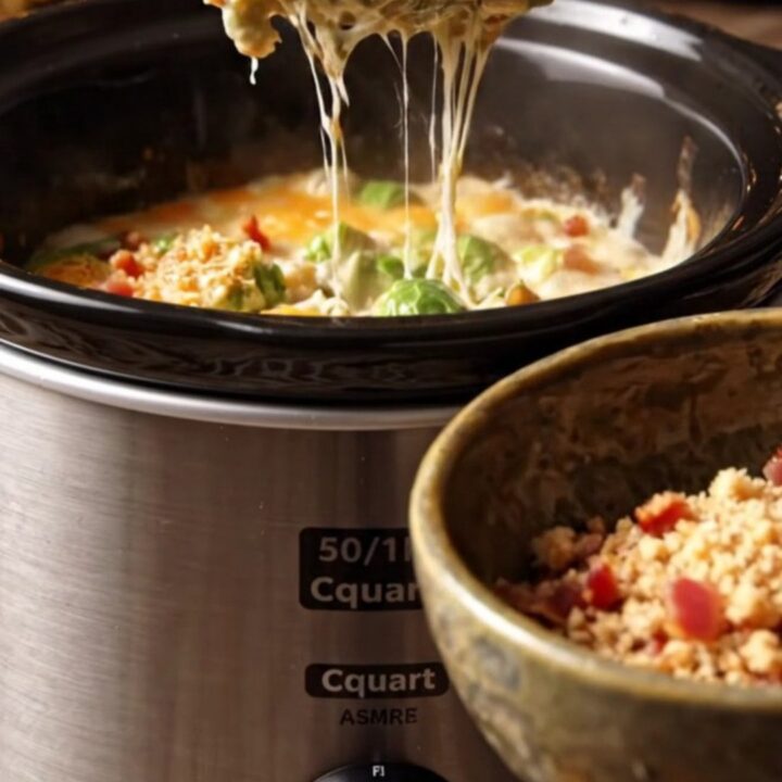 A close-up of a ceramic slow cooker filled with creamy Brussels sprouts au gratin, topped with crispy bacon and breadcrumbs.