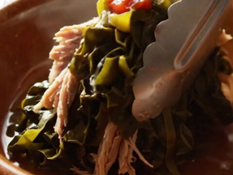 A close-up view of silky, slow-cooked collard greens with shredded smoked turkey in a cream stoneware crockpot.