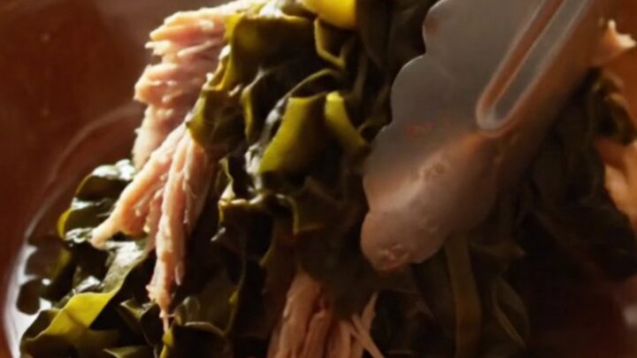 A close-up view of silky, slow-cooked collard greens with shredded smoked turkey in a cream stoneware crockpot.