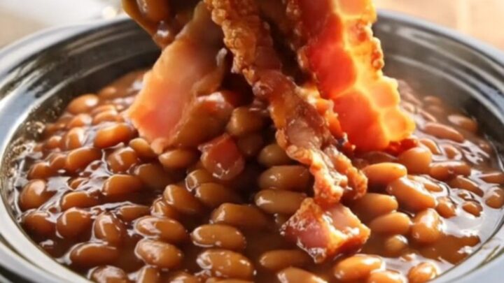 A close-up shot of thick, bubbling maple bacon baked beans in a black ceramic slow cooker.