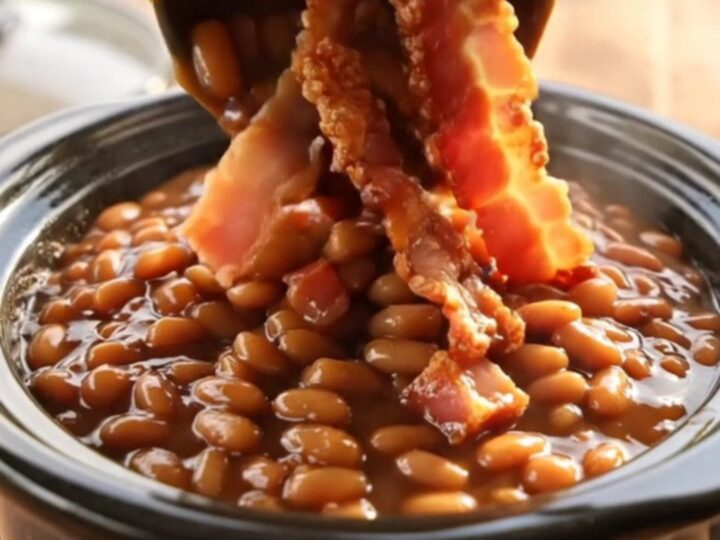 A close-up shot of thick, bubbling maple bacon baked beans in a black ceramic slow cooker.