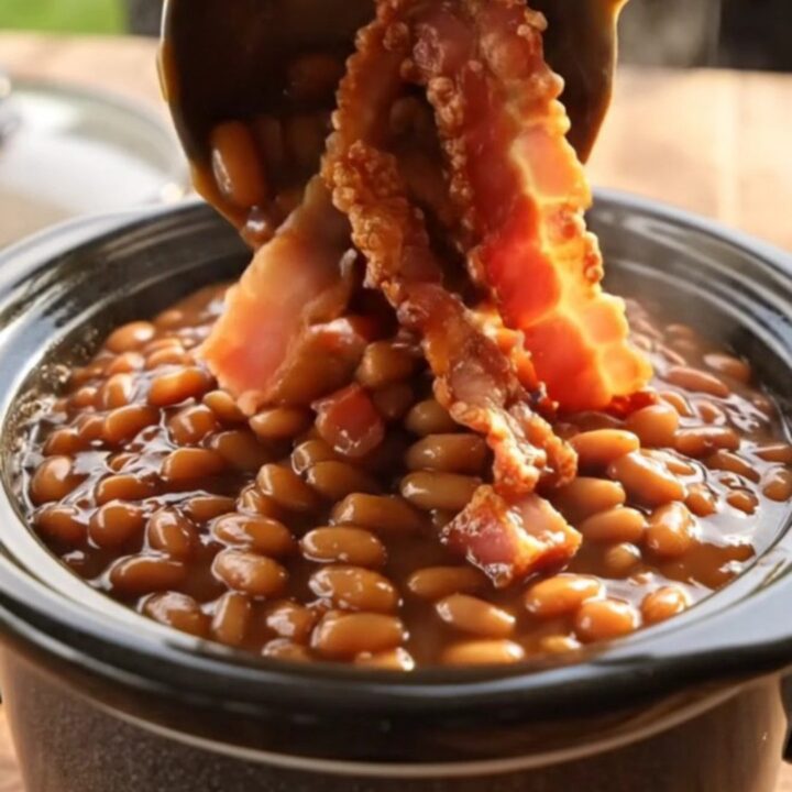 A close-up shot of thick, bubbling maple bacon baked beans in a black ceramic slow cooker.