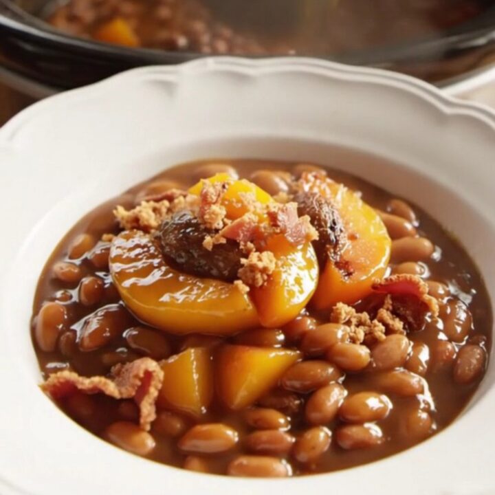 A close-up of a bowl filled with bourbon peach baked beans, garnished with grilled peach slices and crispy bacon.