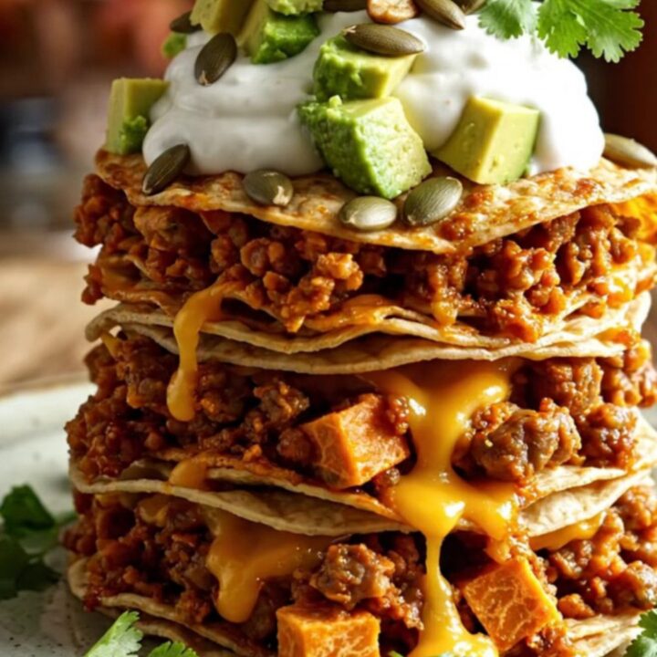 A vibrant stack of slow-cooked enchiladas topped with avocado, sour cream, and cilantro in a rustic kitchen.