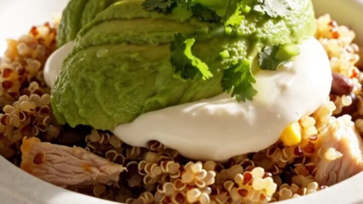 A close-up of a creamy chicken and quinoa burrito bowl topped with avocado, sour cream, and cilantro.