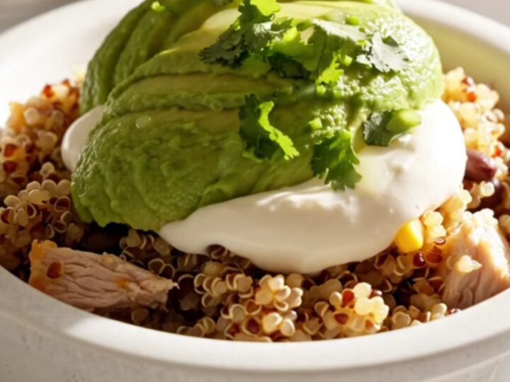 A close-up of a creamy chicken and quinoa burrito bowl topped with avocado, sour cream, and cilantro.