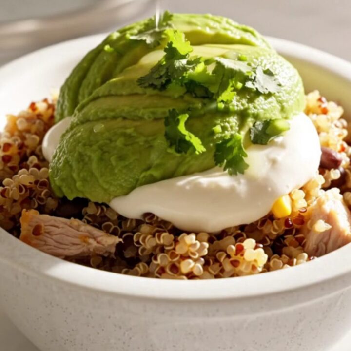 A close-up of a creamy chicken and quinoa burrito bowl topped with avocado, sour cream, and cilantro.