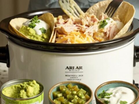 A glossy white slow cooker filled with creamy white queso chicken, with a spoon lifting a portion to show a cheese pull.