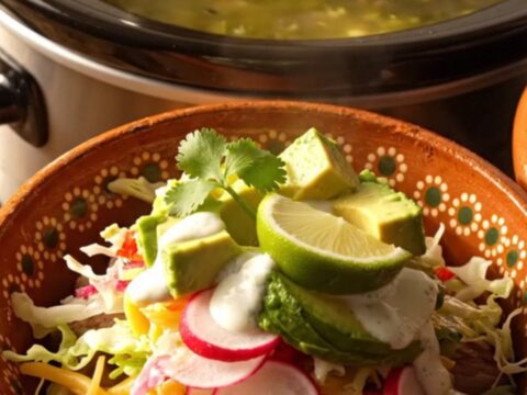 A steaming bowl of green chile pork posole topped with fresh cabbage, avocado, and radishes in a warm kitchen.