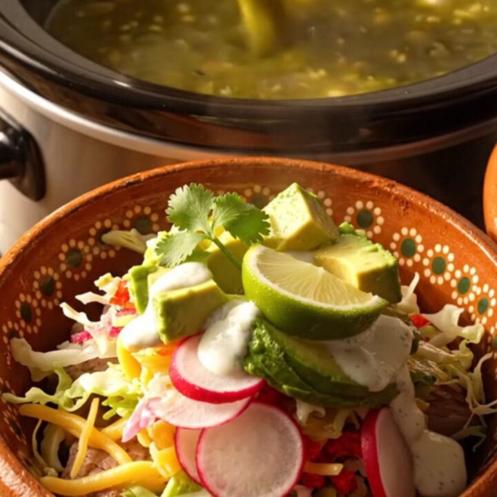 A steaming bowl of green chile pork posole topped with fresh cabbage, avocado, and radishes in a warm kitchen.