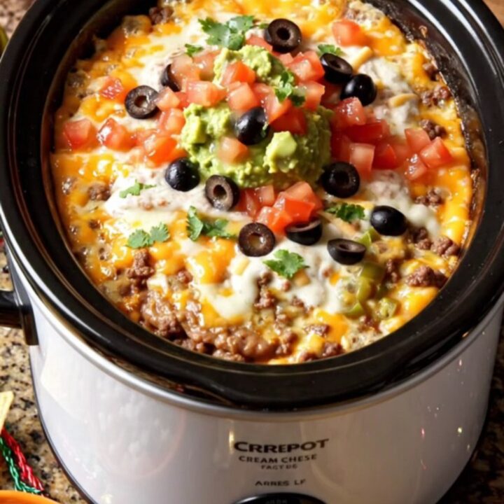 A warm, bubbling seven-layer taco dip in a white oval crockpot, topped with fresh guacamole and cilantro.